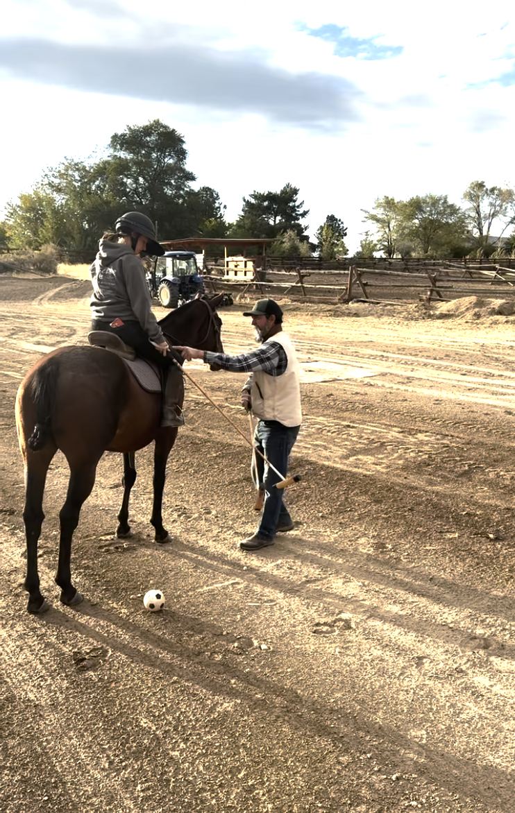 Daniel Fernández, on foot, adjusts a brand-new player's grip on the mallet, with a practice ball on the ground beside the horse.