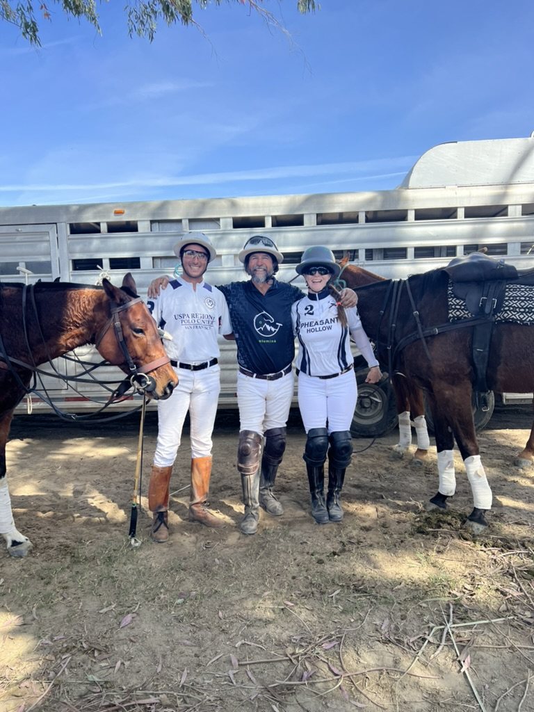 Facundo, Daniel Fernández and Rebecca Clark stand arm in arm in polo whites beside two horses and a trailer.